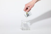 Pouring BRDY JUICE electrolyte powder from a sachet into a glass of clear water — step 1 of the mix